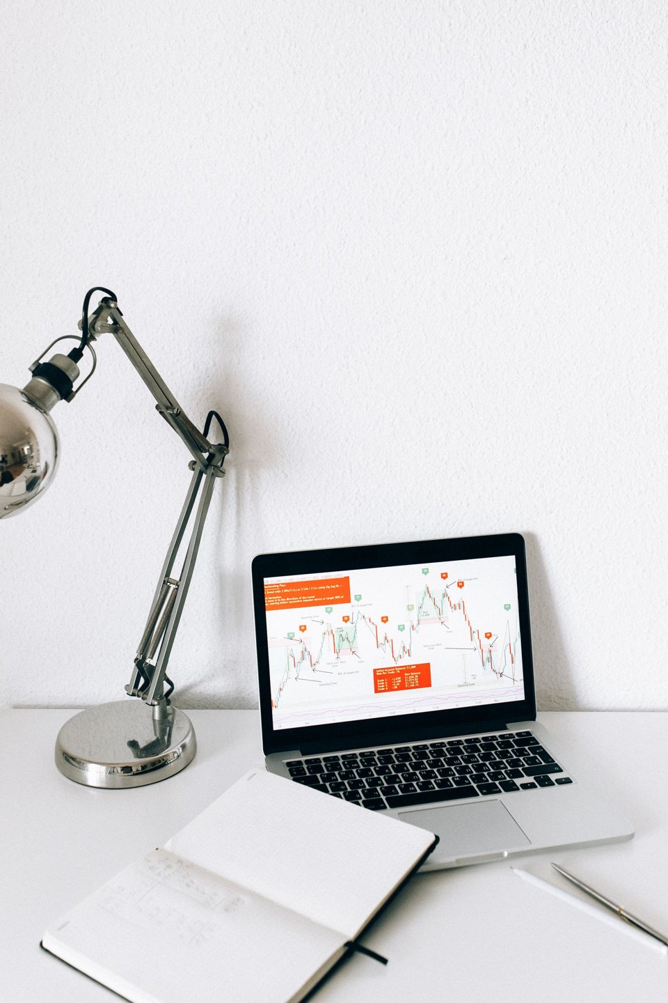 A laptop displaying a line chart beside an open notebook and desk lamp.